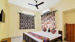 A bedroom with a bed, curtains, a ceiling fan, an air conditioning unit, a wooden wardrobe, and a small table with a remote control. - Fabhotel Florence Palace (Kolkata)