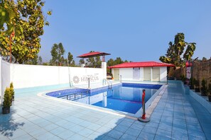A swimming pool area with a red-roofed building, a white wall with a logo, and a clear blue sky. - Fabhotel Wild Hibiscus Resort (Ramnagar)