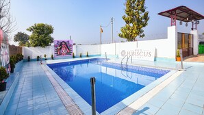 A swimming pool with a clear blue water, surrounded by a white wall and a red-roofed structure. - Fabhotel Wild Hibiscus Resort (Ramnagar)