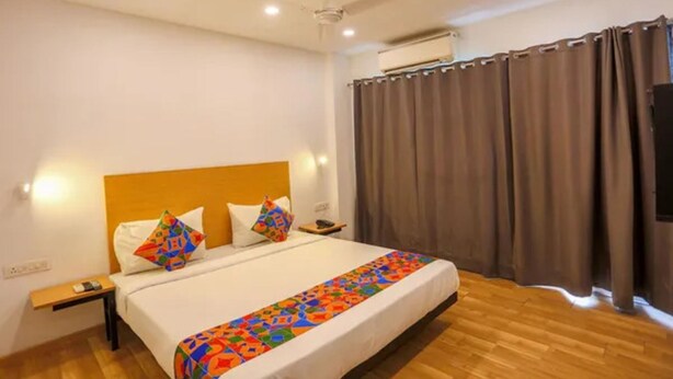 A hotel room with a wooden floor, a bed with a colorful blanket, a headboard, a nightstand, a phone, and a window with curtains. - Fabhotel Bellmore Stay & Homes (Gurugram)