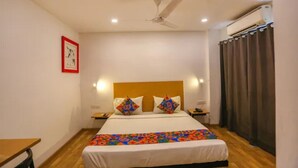 A hotel room with a bed, a ceiling fan, a nightstand, a bedside table, and a window with curtains. - Fabhotel Bellmore Stay & Homes (Gurugram)