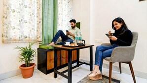 Two people sitting on chairs, one on a couch and one on a table with a tray of food and a cup. - Via Rajdanga Retreat (Kolkata)