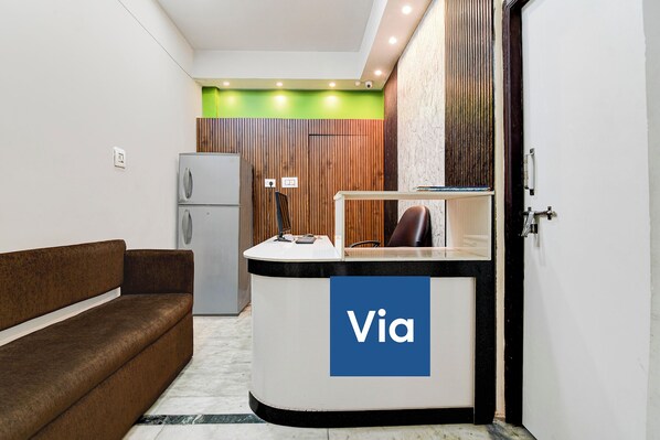 A reception desk with a 'Via' sign, a brown sofa, a refrigerator, and a wooden wall. - Via Rajdanga Retreat (Kolkata)