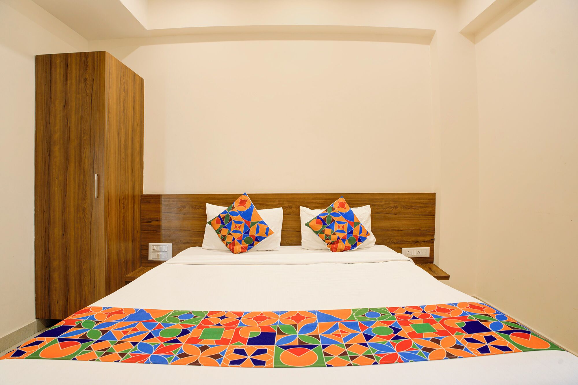 A hotel room with a wooden wardrobe, a bed with a colorful geometric-patterned bedspread, and two pillows with geometric designs.