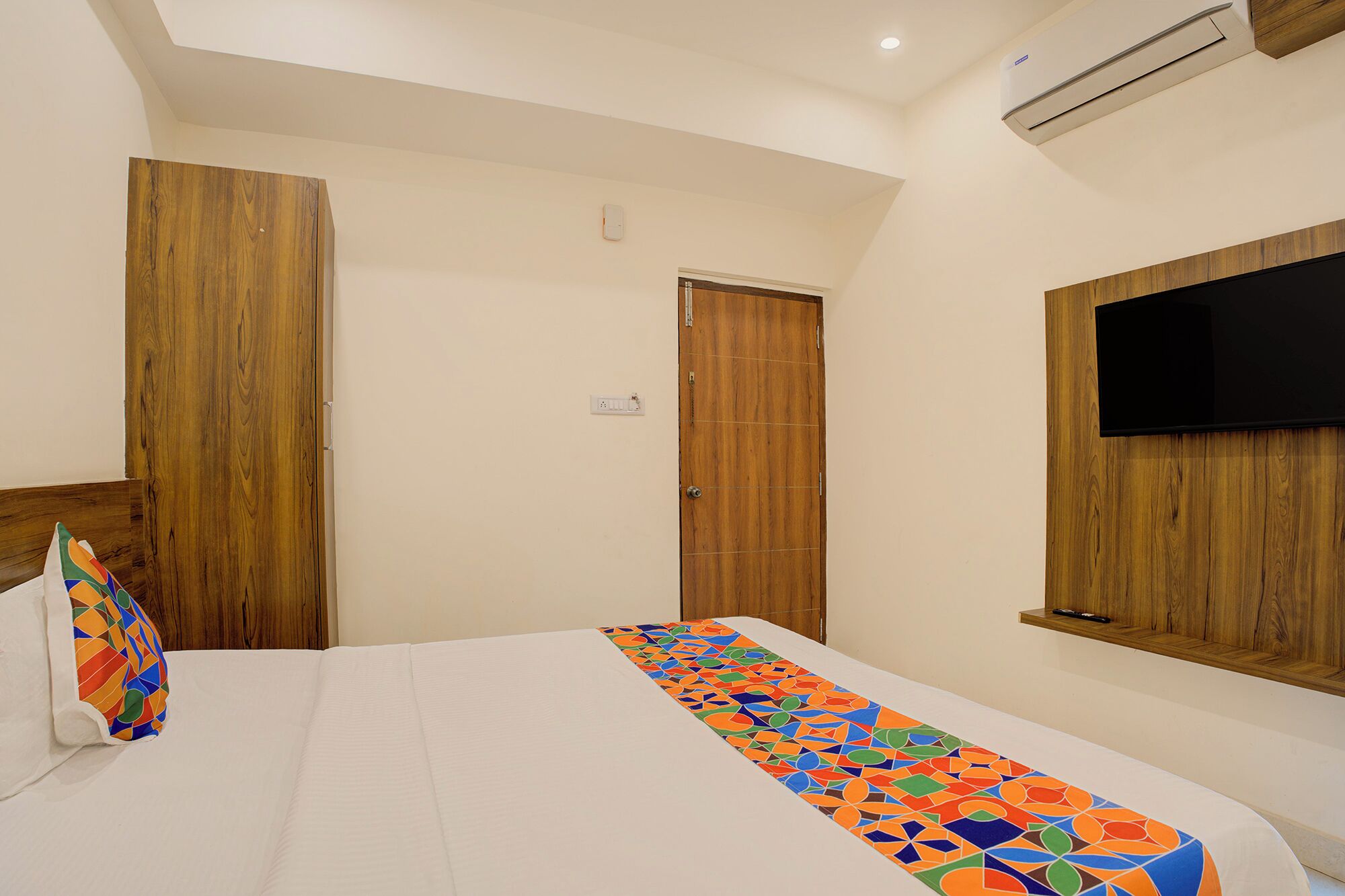 A bedroom with a bed, wooden wardrobe, a television mounted on the wall, and a ceiling fan.