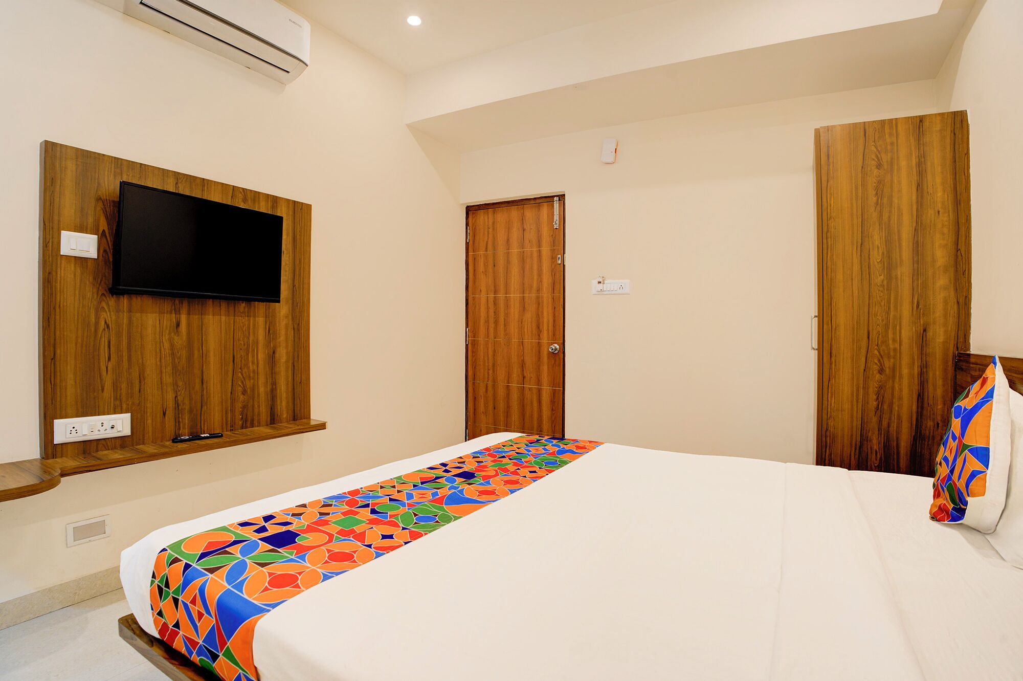 A hotel room with a bed, a television mounted on a wooden panel, a wooden wardrobe, and a door.