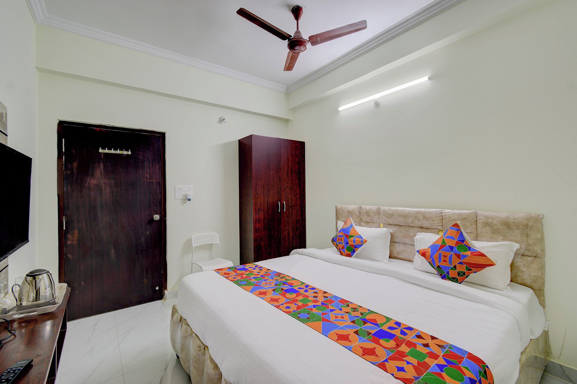 A hotel room with a bed, a ceiling fan, a desk with a kettle, and a TV.