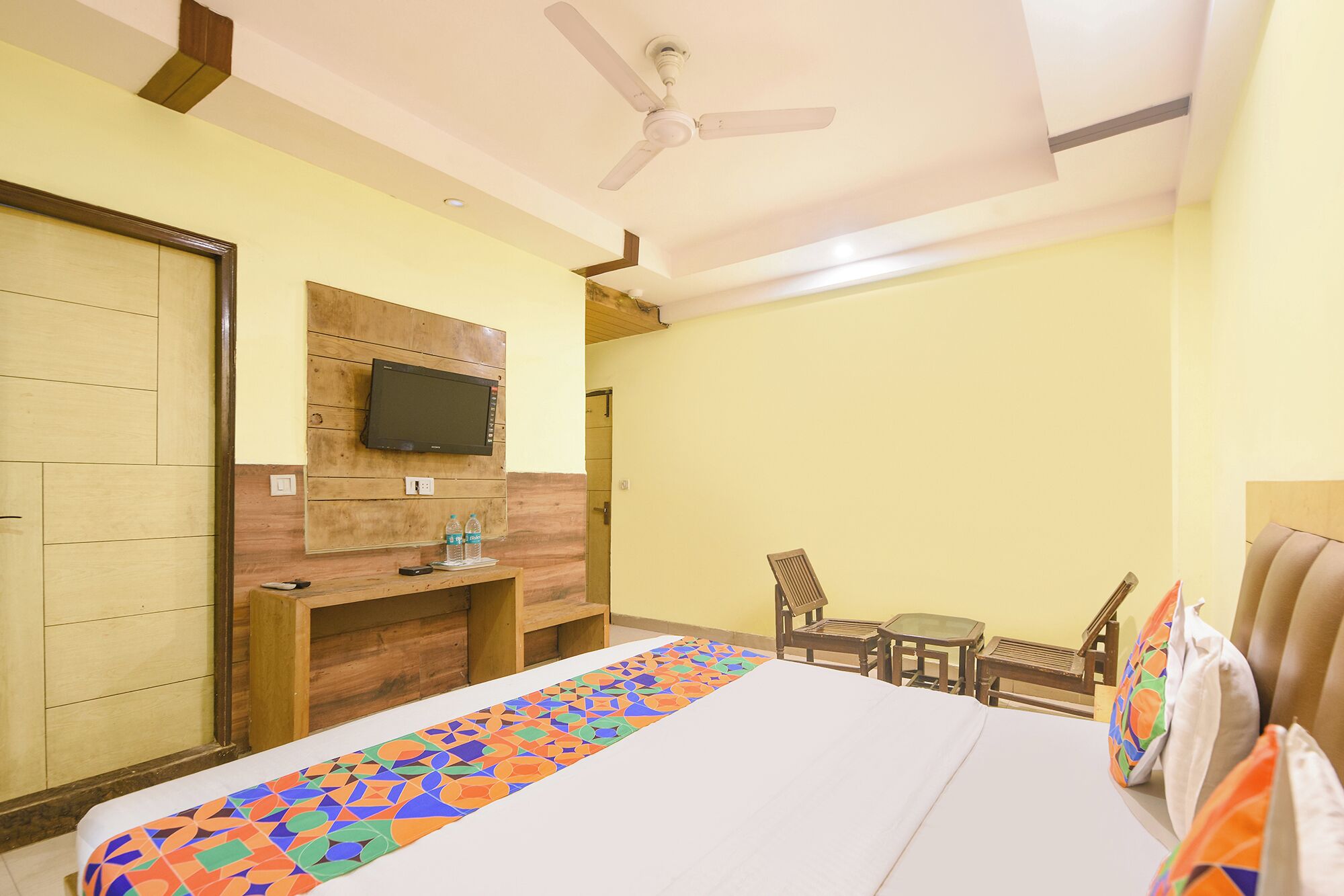 A hotel room with a bed, a TV mounted on a wooden panel, a ceiling fan, and a small table with chairs.