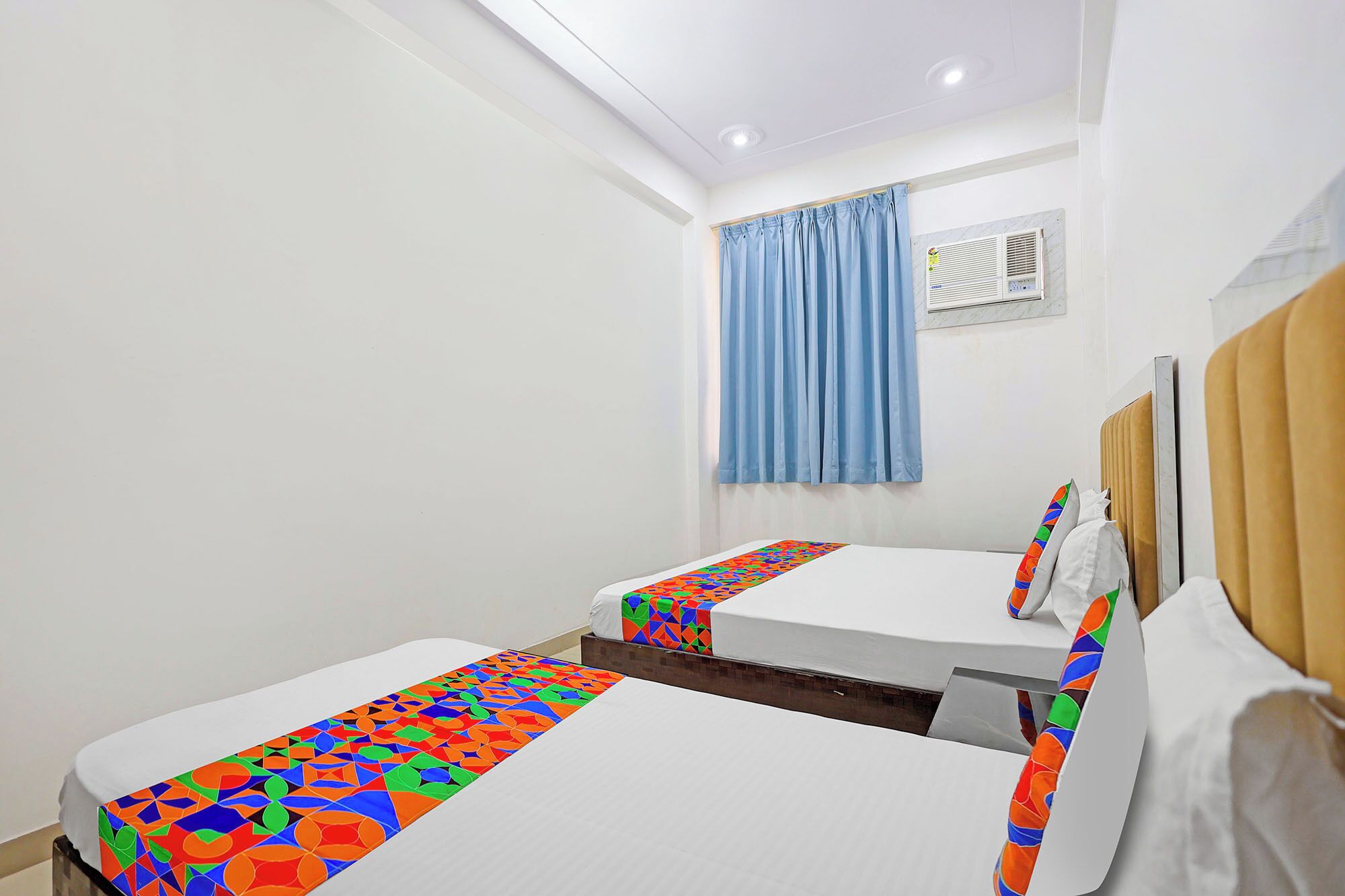 A hotel room with two beds, a window with curtains, a ceiling light, and a wall-mounted air conditioning unit.