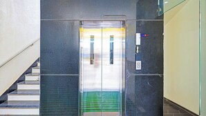 An elevator with a glass door, a control panel with buttons, and a reflection of a staircase. - Fabhotel Shri Hari Singh Dham (Vrindavan)