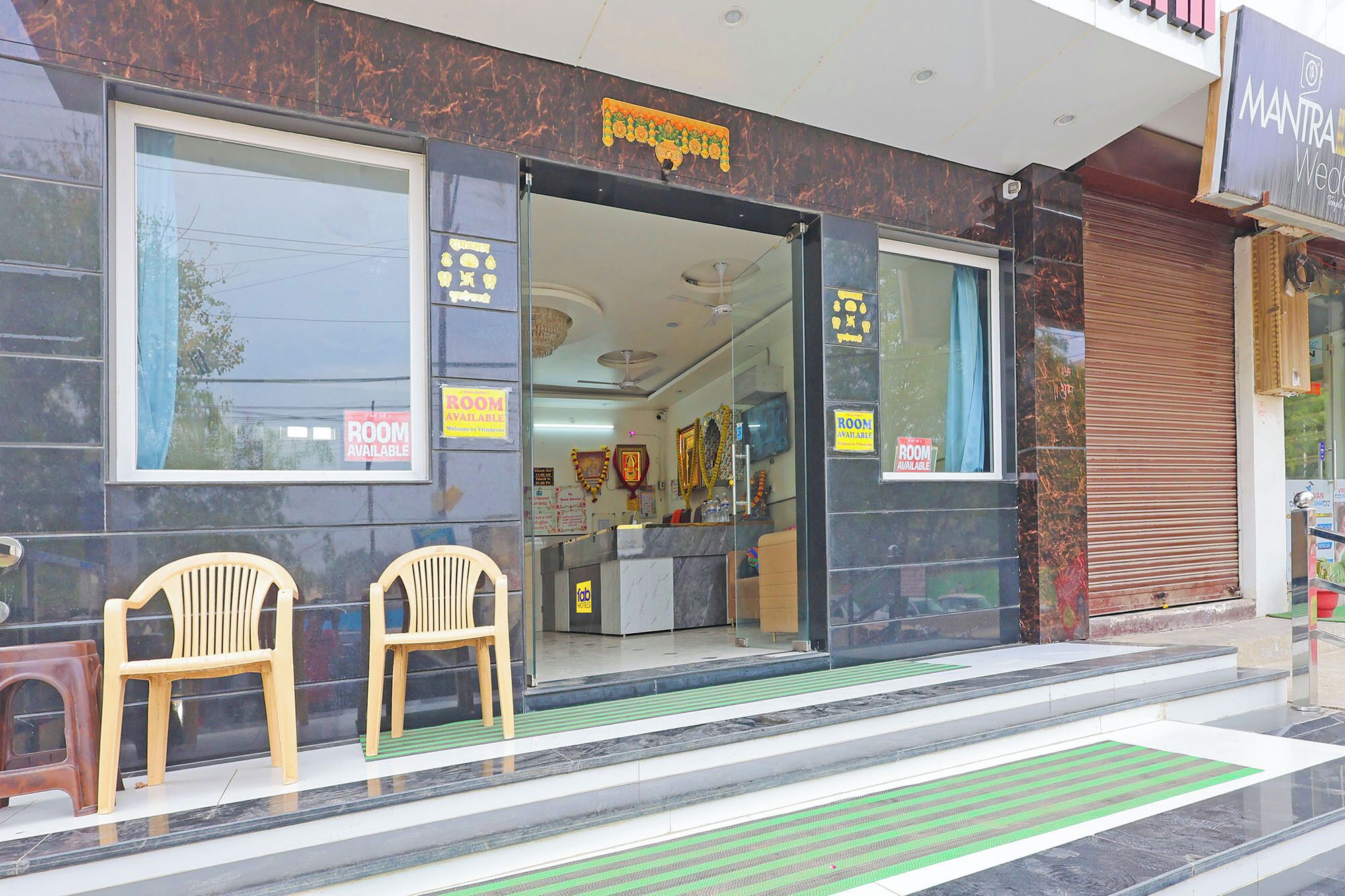 A hotel entrance with a sign indicating rooms are available, a reception desk inside, and two wooden chairs outside.