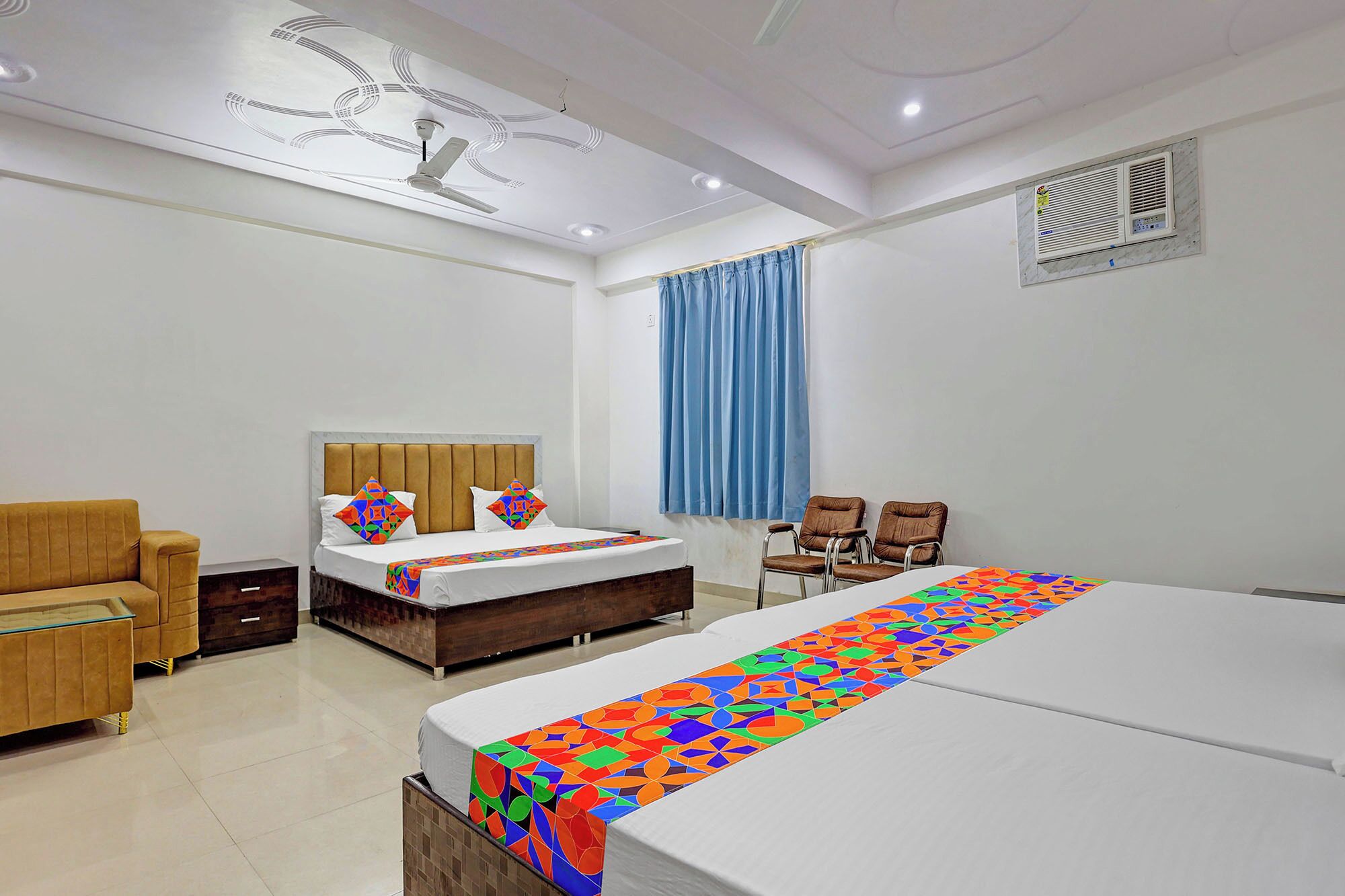 A hotel room with a large bed, a smaller bed, a sofa, two chairs, a ceiling fan, and a window with curtains.
