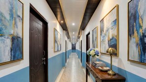 A hallway with blue and white walls, a marble floor, and framed artwork. There is a wooden door and a table with a lamp and flowers. - Via Atithi Inn (Varanasi)