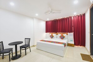 A hotel room with a bed, a small table with two chairs, a ceiling fan, and a window with red curtains. - Fabhotel Happy Stay (Noida)