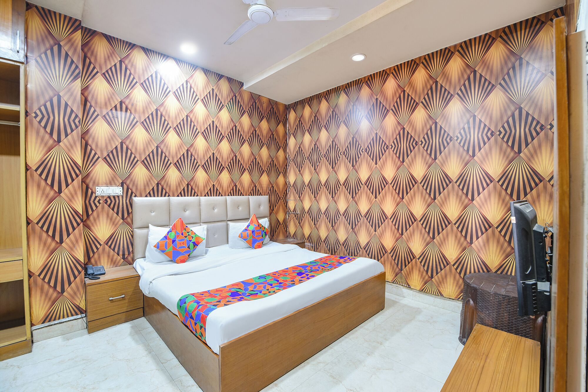 A bedroom with a patterned wallpaper, a bed with a colorful geometric throw, a bedside table, and a television.