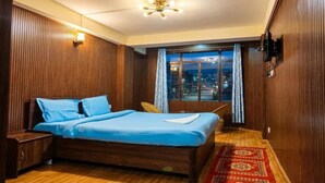 A bedroom with a wooden bed, a nightstand, a window with a view of the city at night, and a patterned rug. - Fabhotel Lorung Residency (Darjeeling)