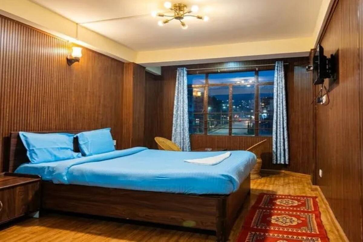 A room with a bed, a nightstand, a window with curtains, a wooden floor, and a ceiling light fixture.