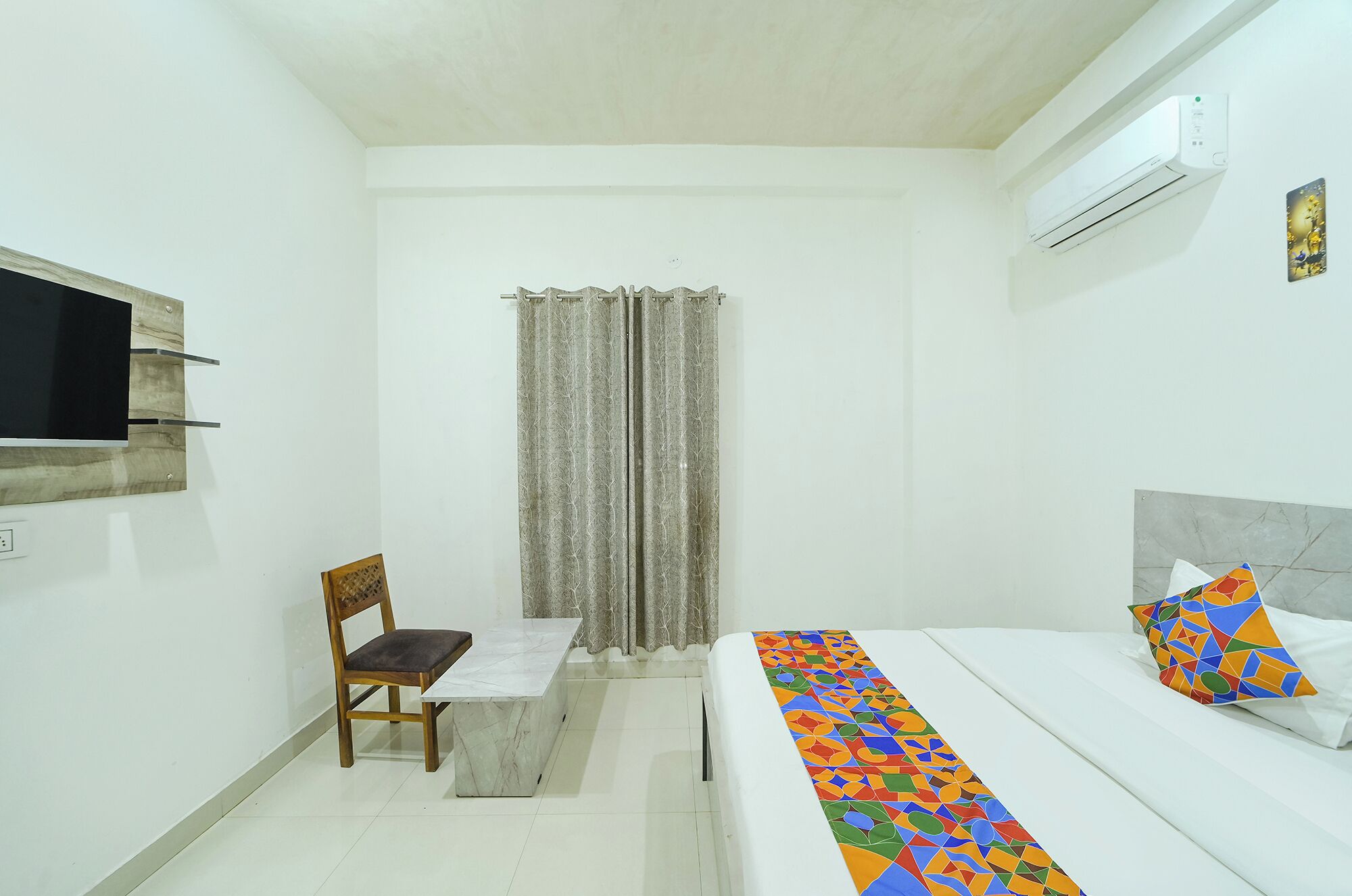 Una habitación pequeña y limpia con una cama, una silla, un televisor montado en la pared y un aire acondicionado.