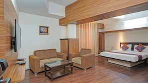 Habitació d'hotel moderna amb sòl de fusta, un sofà, una cadira, un llit amb almoadins de colors, una televisió de pantalla plana i un quadre a la paret.