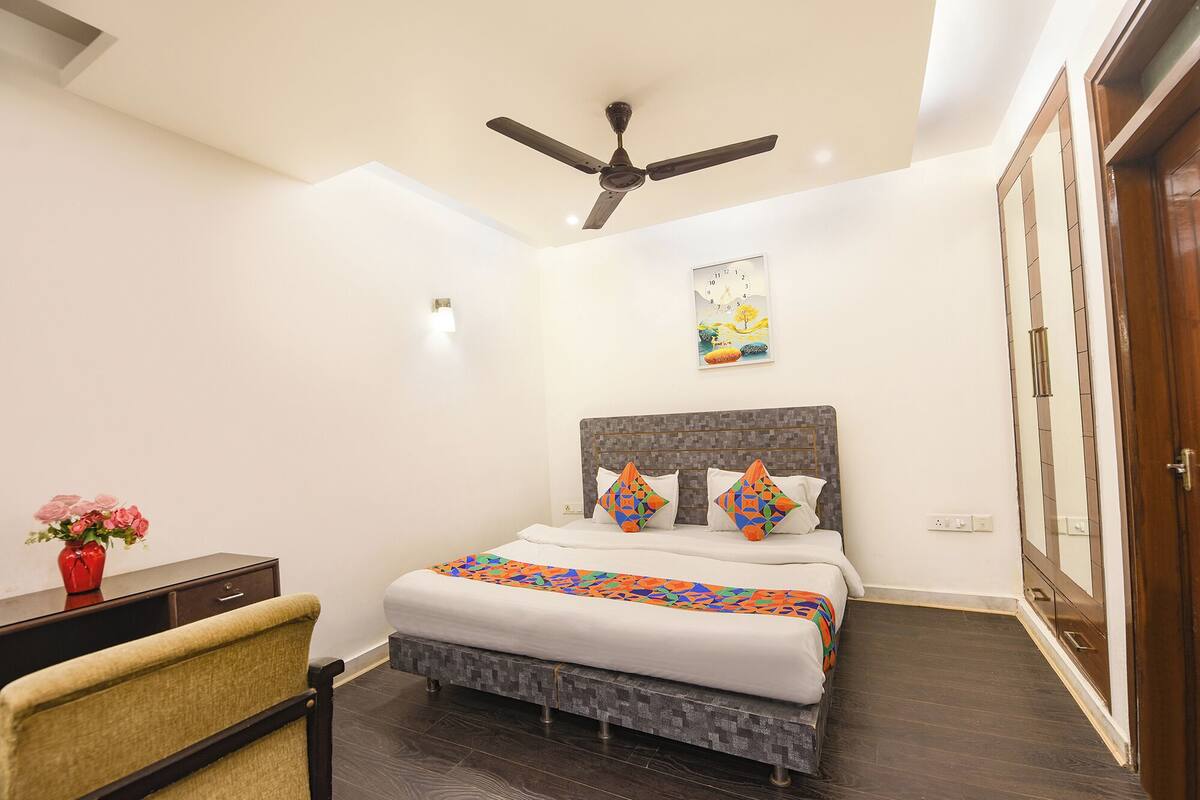 A bedroom with a bed, a ceiling fan, a nightstand, a chair, a vase with flowers, and a painting on the wall.