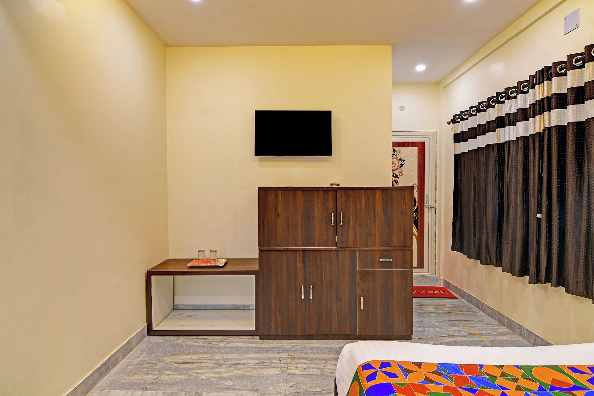 Habitación de hotel con una cama, un armario de madera, una mesita con bandeja, un televisor de pantalla plana en la pared y una cortina estampada.