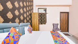 A hotel room with a large bed, a blue upholstered headboard, patterned wallpaper, a wooden door, and a grey upholstered chair. - Via Town Plaza (New Delhi)