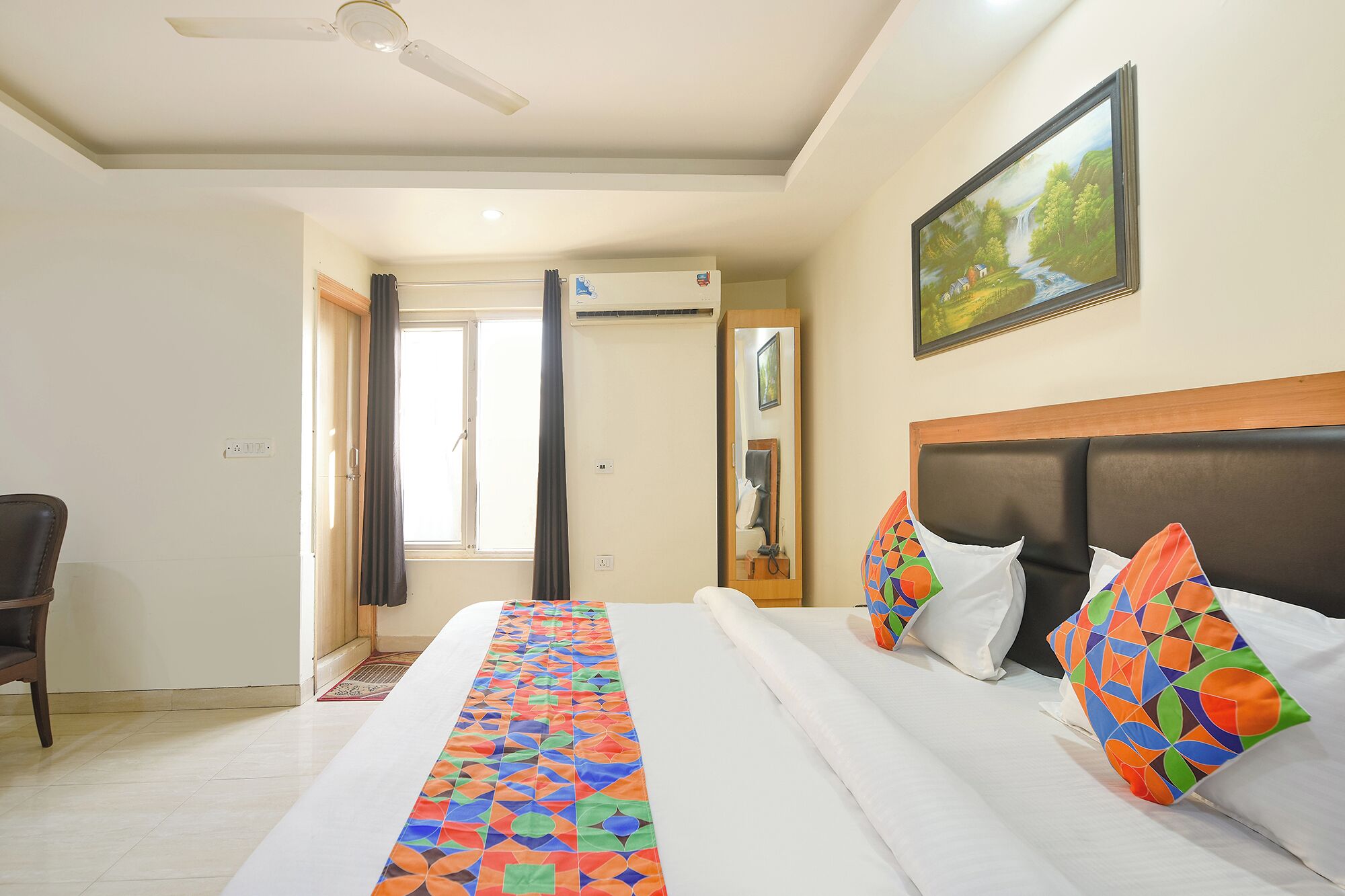 A hotel room with a large bed, a headboard, a painting on the wall, and a window with curtains.