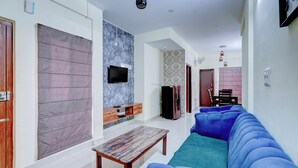 A living room with a blue sofa, a wooden coffee table, a flat-screen TV, and a refrigerator. - Fabhotel Address 24 Suites (Bengaluru)