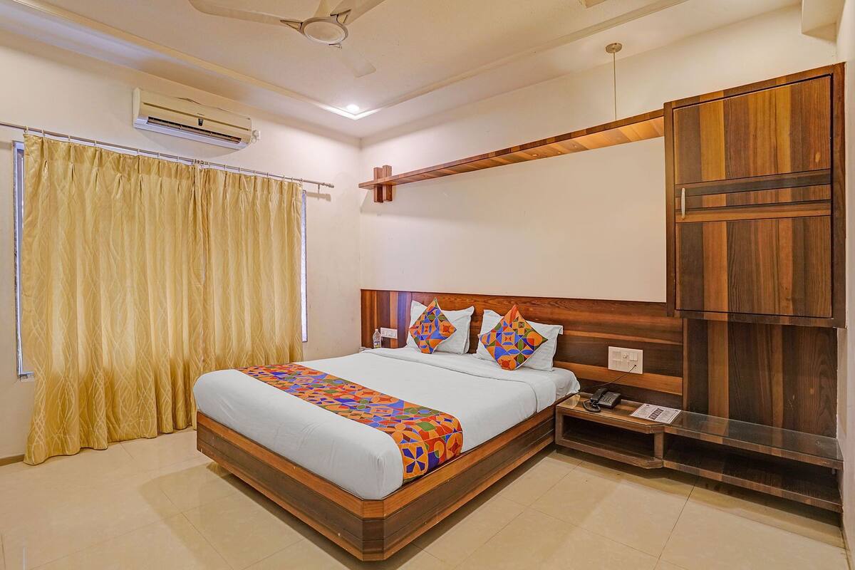 A bedroom with a bed, curtains, an air conditioning unit, a wooden headboard, a bedside table with a phone, and a wall-mounted shelf.