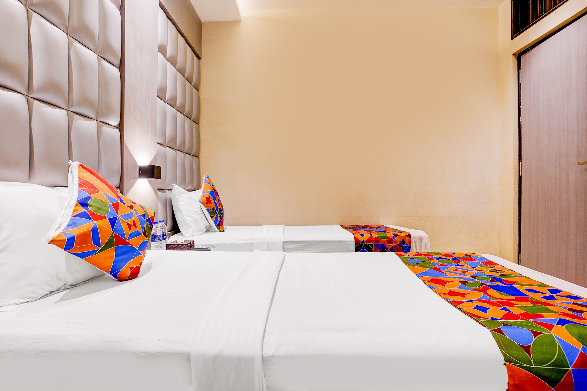 Habitación de hotel con dos camas, cada una con una colcha blanca y almohadas de colores con motivos geométricos. La habitación tiene una pared color beige y una puerta de madera.