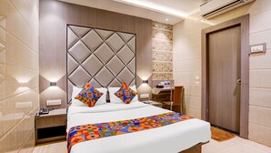 A hotel room with a large bed, a wooden headboard, a nightstand, a bedside table, a chair, and an air conditioning unit. - Fabhotel Abrol Suites (Mumbai)