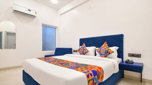Quarto de hotel com uma cama grande, cabeceira azul, manta colorida, mesa de cabeceira com telefone e janela com persianas.