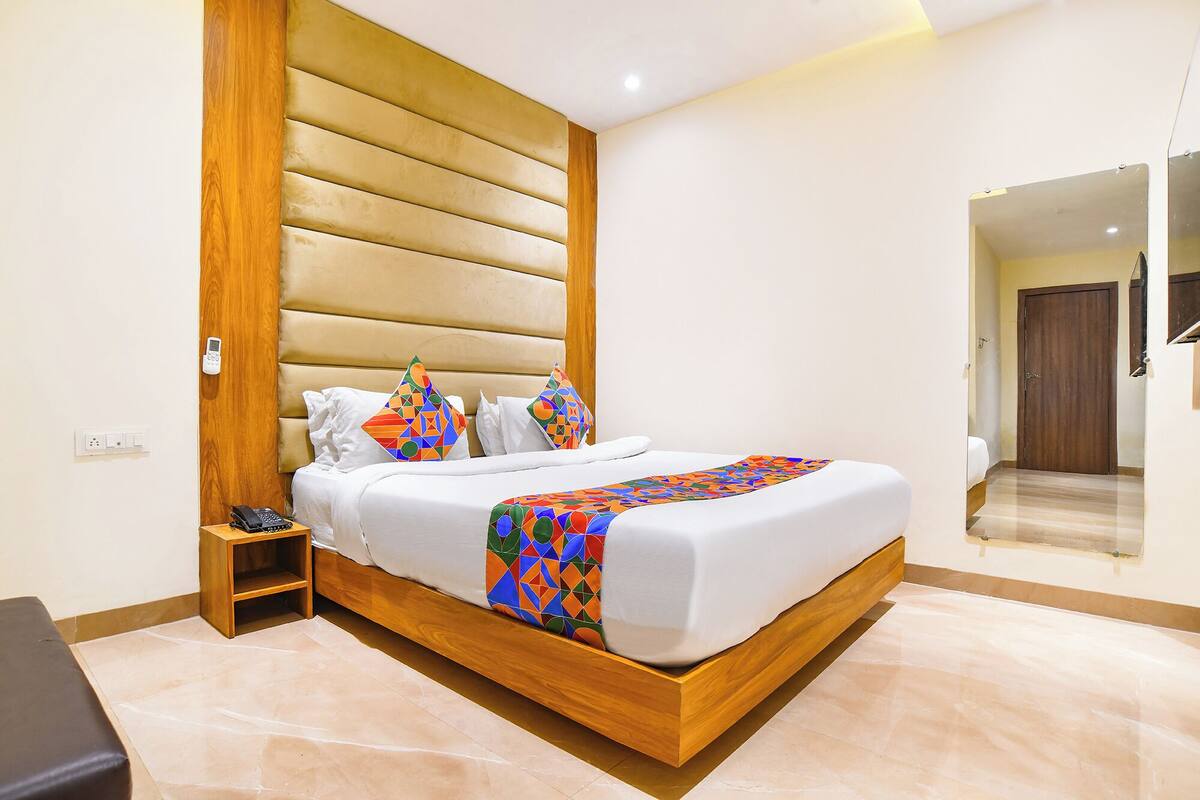 A modern hotel room with a wooden headboard, a large bed with white bedding, colorful geometric throw pillows, a bedside table with a phone, and a mirror on the wall.