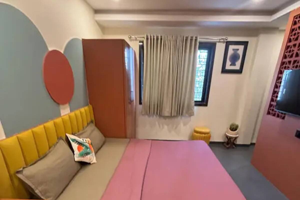 A bedroom with a bed, a ceiling fan, a window with curtains, a small table, and a TV mounted on the wall.