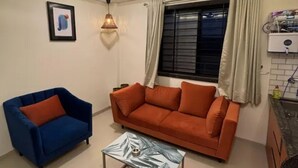 Uma sala de estar com ventilador de teto, uma poltrona azul, um sofá laranja e uma mesa de centro estampada.