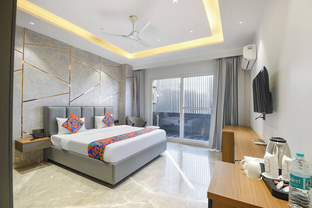 A modern hotel room with a large bed, a desk with a coffee maker, a flat-screen TV, and a balcony with curtains.