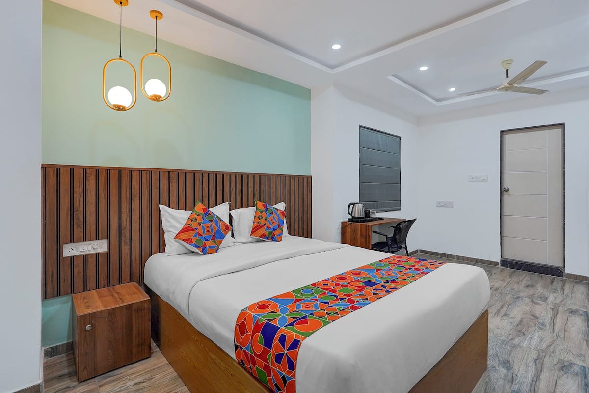 A modern hotel room with a wooden headboard, a bed with colorful geometric pillows, a desk with a chair, a ceiling fan, and a large window.