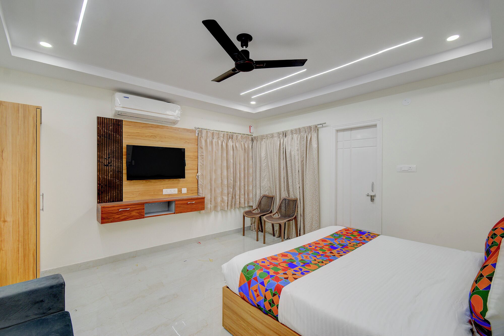 A modern hotel room with a bed, a TV mounted on the wall, a ceiling fan, and a window with curtains.