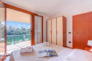 A hotel room with a bed, a large window, a wardrobe, and a view of a lake. - Albarella S4 1, Isola di Albarella, Italy (Isola di Albarella)
