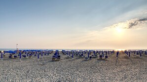 Una playa con hileras de sombrillas azules y sillas de playa, un cielo despejado y una puesta de sol a lo lejos.