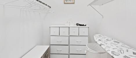 A compact, white room with a built-in wardrobe, a small table with a trash bin, and a wall-mounted drying rack.