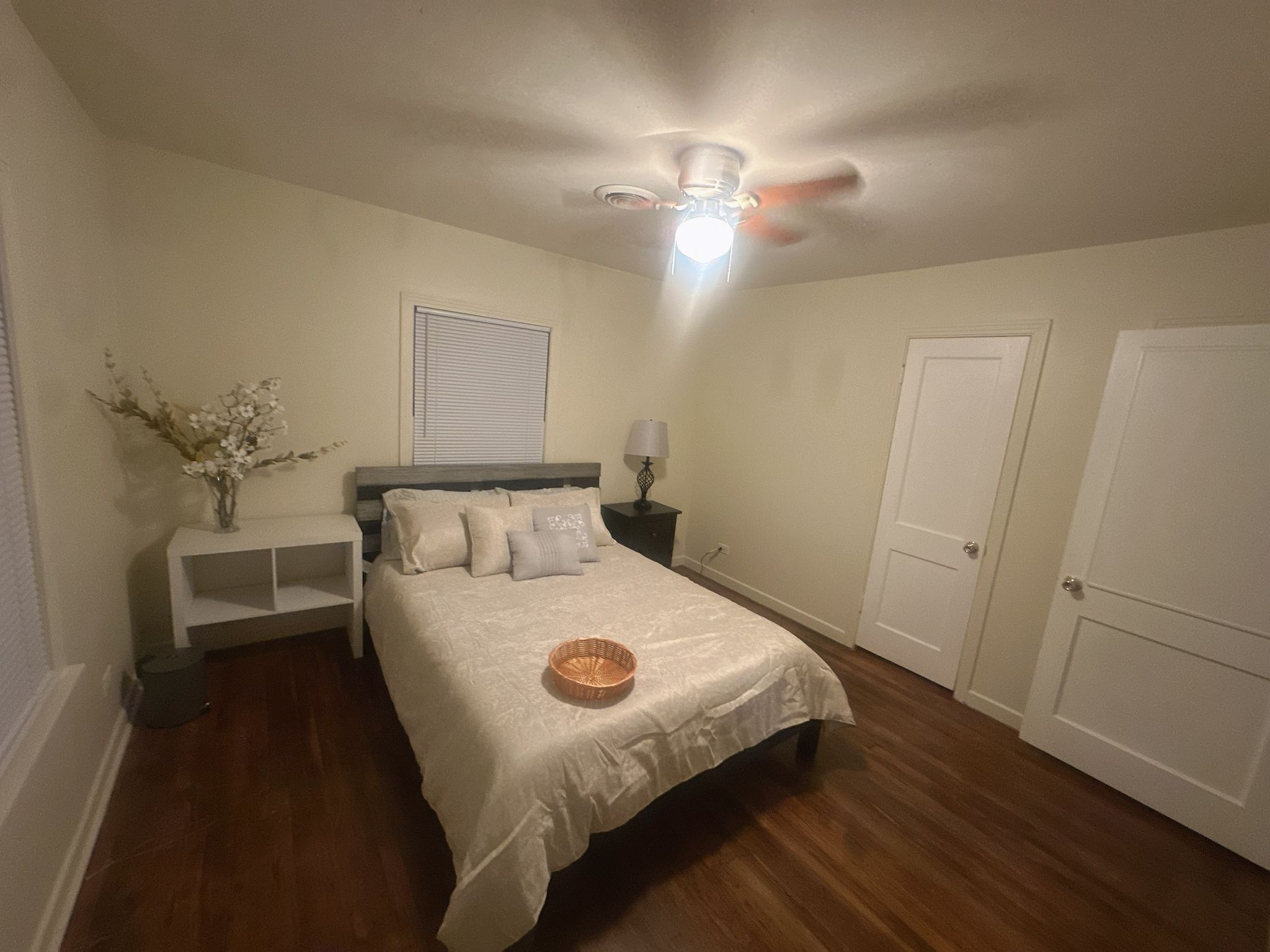A bedroom with a bed, nightstands, a ceiling fan, a window with blinds, and a door.