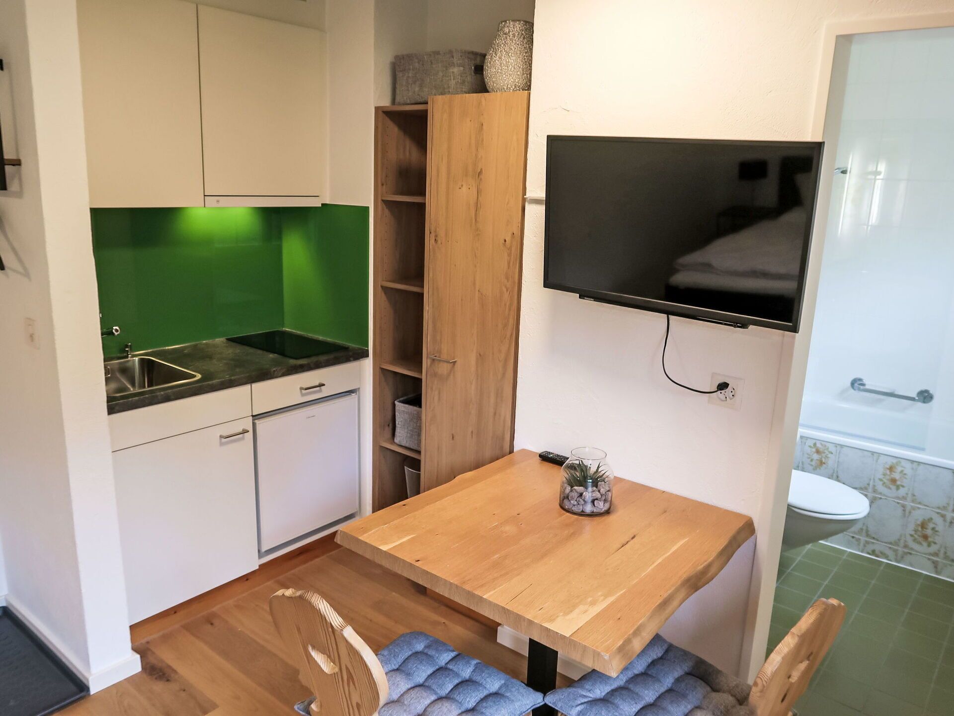 Cocina compacta con un lavamanos, un refrigerador pequeño y un armario de madera. Hay un televisor montado en la pared sobre una mesa de madera con un florero de vidrio.