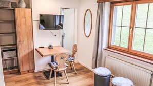Ein gemütlicher Raum mit Holztisch, Wandfernseher, Fenster mit Vorhängen und Heizkörper.