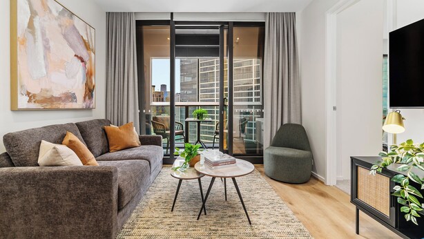 A modern living room with a grey sofa, a round coffee table, and a grey armchair. A large abstract painting is on the wall, and a TV is mounted on the wall. - Modern 1BR Prima Tower • Walk to Crown (Melbourne)