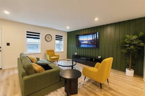 Un salon moderne avec un canapé vert, des fauteuils jaunes, une table basse, un téléviseur fixé au mur et une plante en pot.
