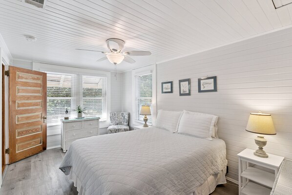 A bedroom with a large bed, a ceiling fan, a dresser, a chair, and a bedside table with a lamp. - NEW LISTING! Seabreeze Cottage | Charming St. Simons Island Beach Cottage Near Pier Village (Saint Simons Island)