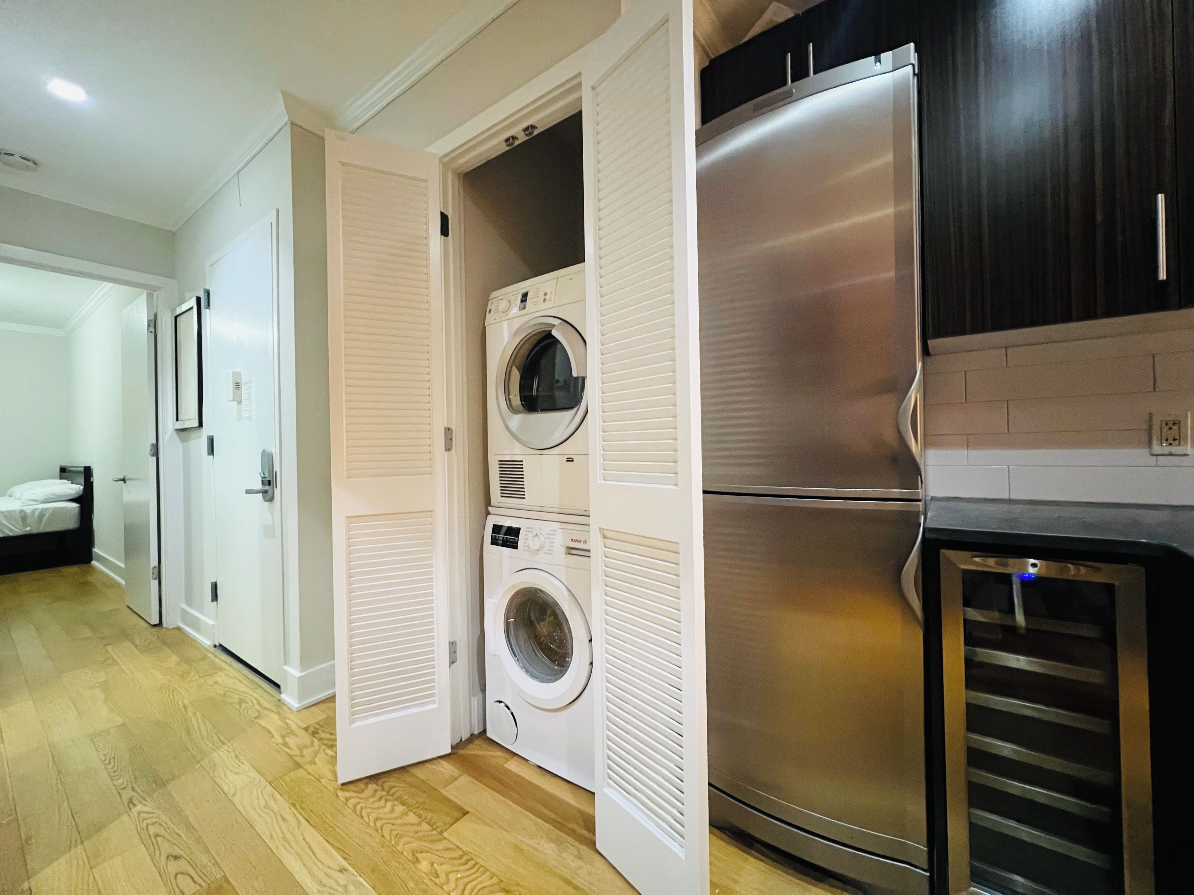 A laundry room with stacked washers and dryers, a stainless steel refrigerator, and a wooden floor.