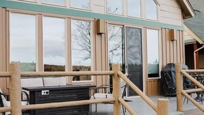 Un edificio moderno con amplios ventanales, un área de estar al aire libre techada con mesas y sillas, y una escalera de madera que conduce a la entrada.