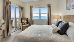 Een slaapkamer met een groot bed, een stoel met een kussen, een lamp en uitzicht op zee door een raam.